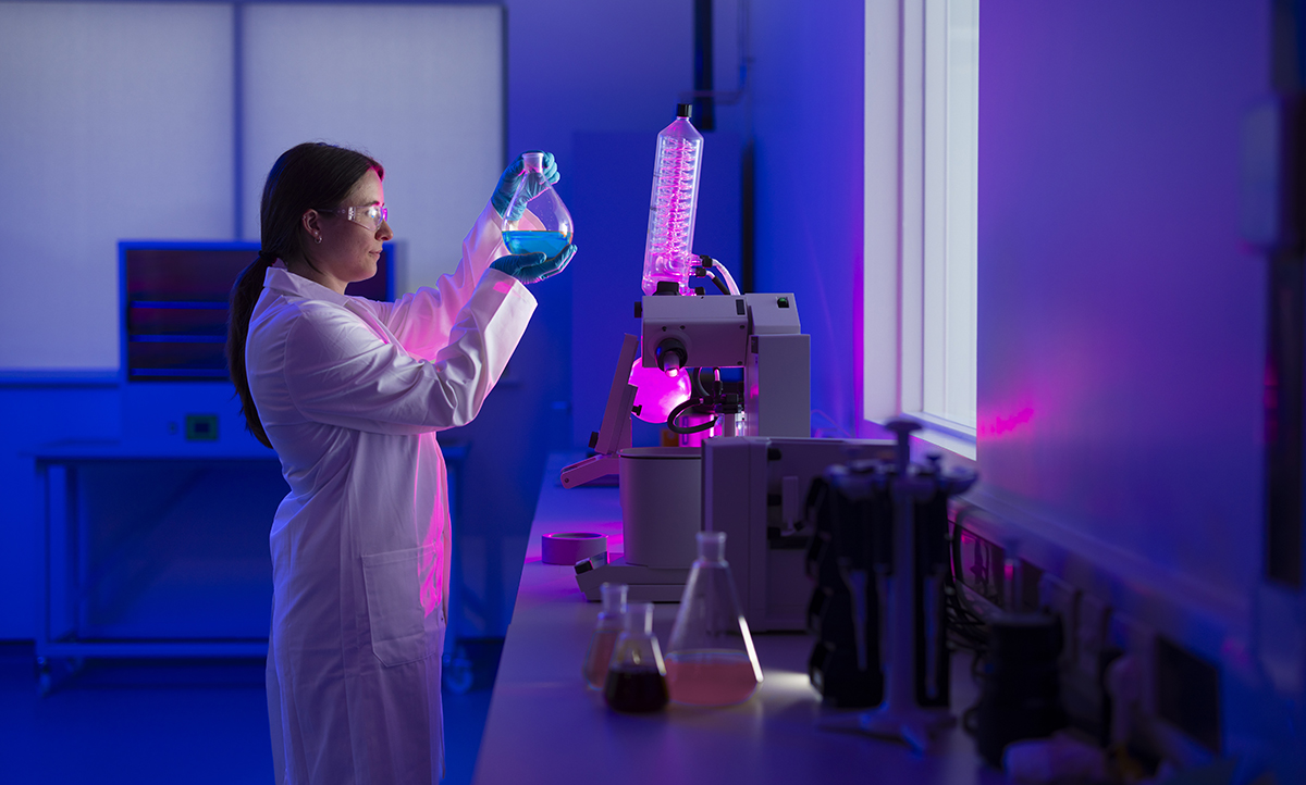 A woman in a lab coat in a purple back-lit lab holding a flask filled with blue liquid