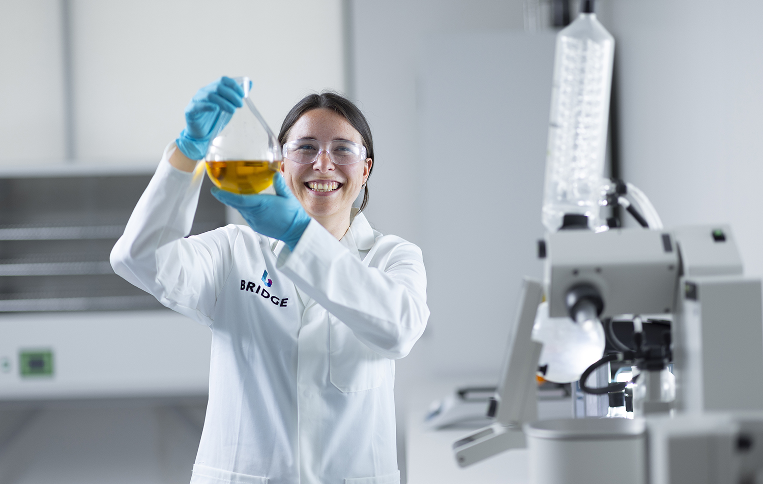 A smiling woman in a lab coat holding a flask of liquid