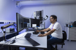 A man sat in front of computer screens with a FIB-SEM in the background